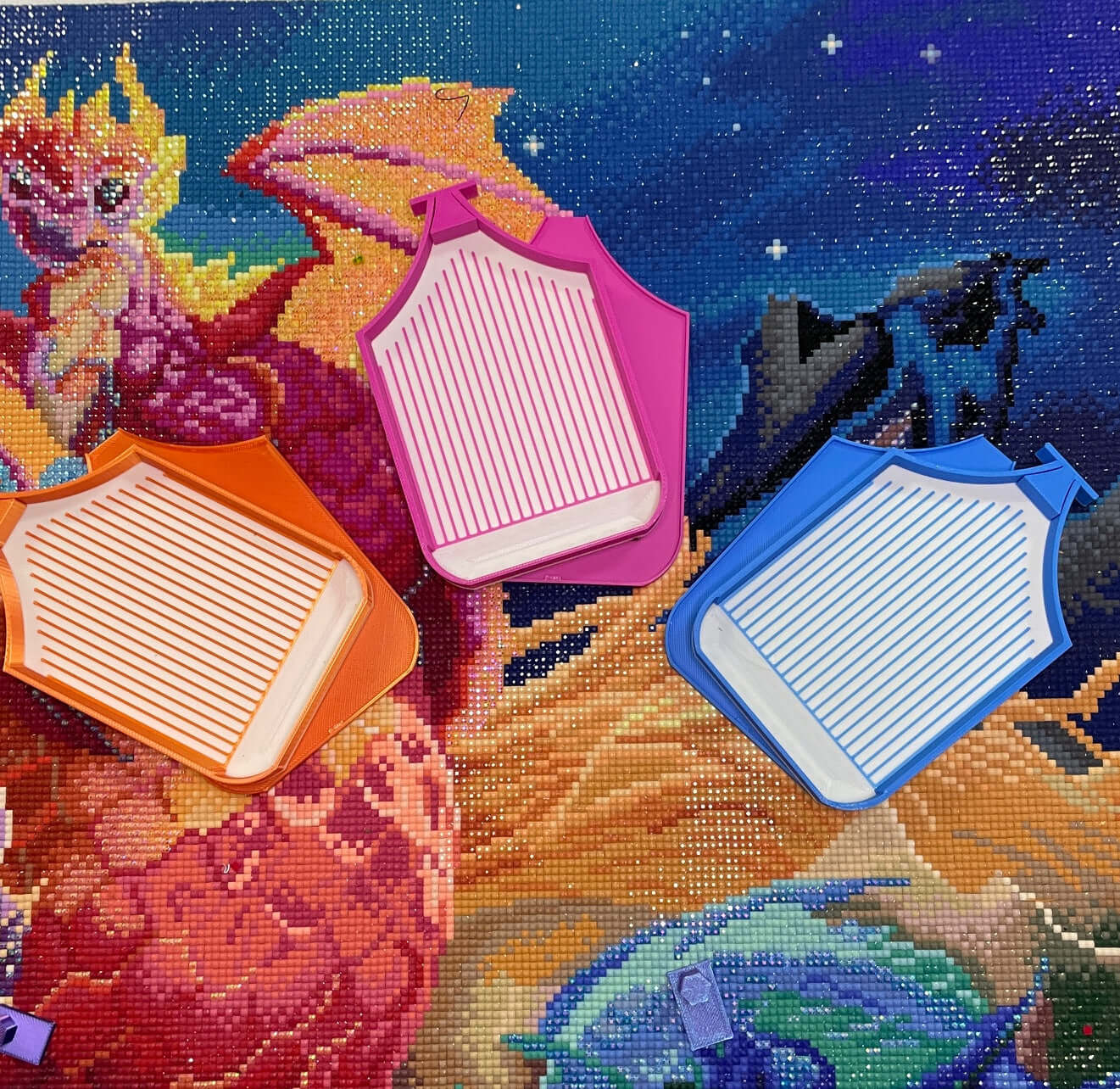 Three Dragonling Trays with white bases in orange, pink, and blue colors displayed on a dragon-themed diamond art background.